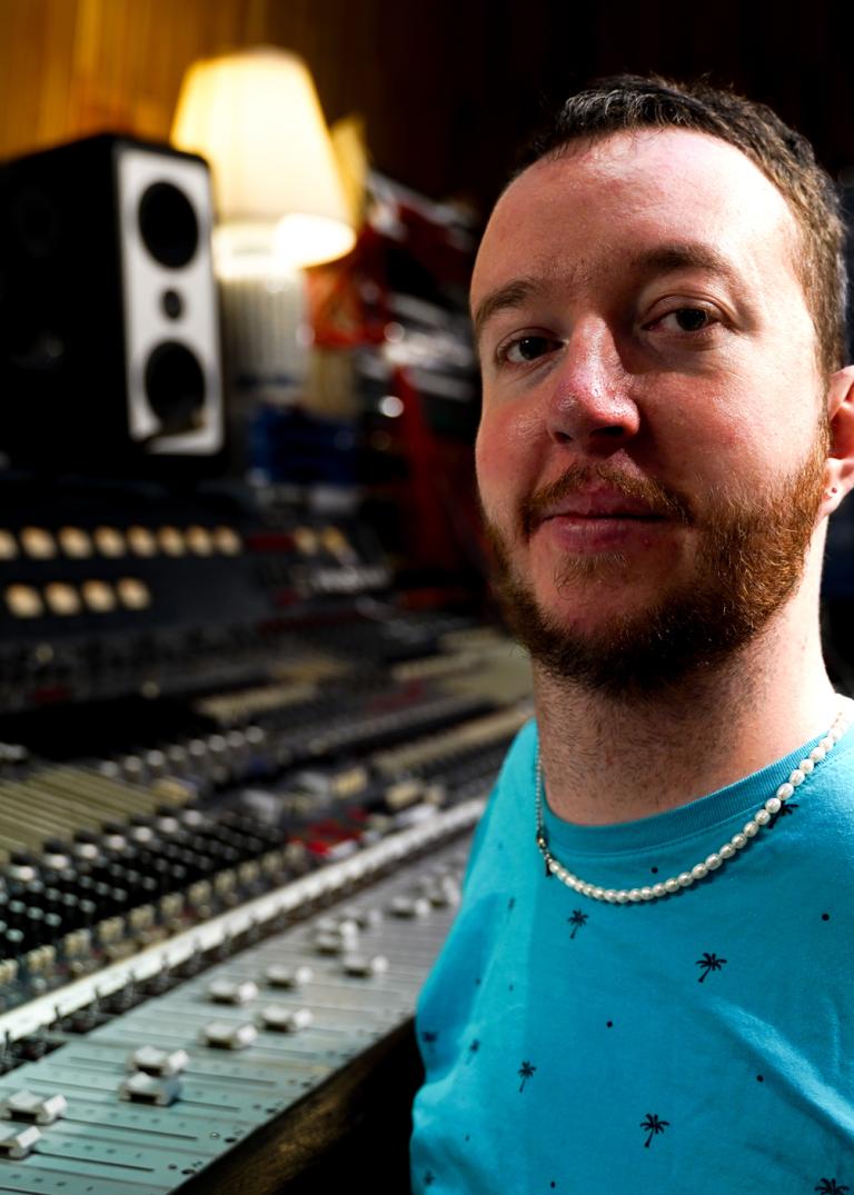 Kompositören och musikern Tim Palm med rött skägg och turkos tshirt sitter framför ett stort mixerbord.
