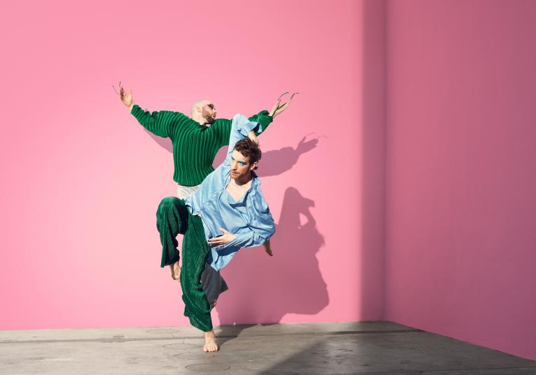 Två dansare i ljusblå och skogsgröna fladdriga kläder dansar yvigt och luftigt framför en rosa vägg.