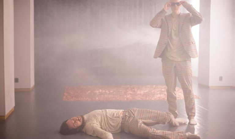 En dansare ligger på golvet vid fötterna av en annan dansare i kostm som sätter på sig solglasögon i ett rökfyllt rum.