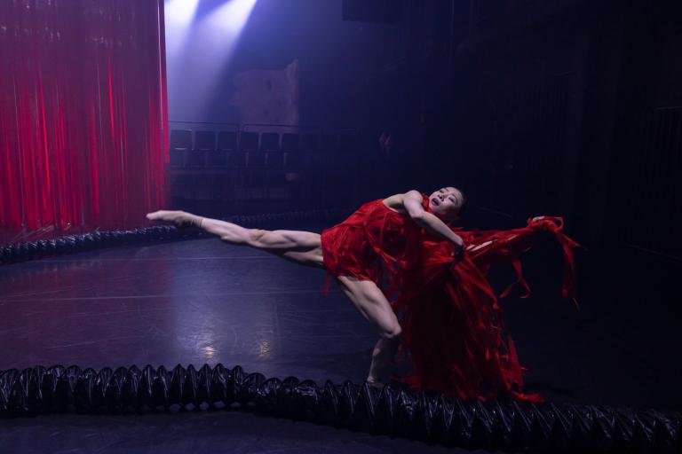 Dansaren Jing Yi Wang i klarröd klänning utav fransar står på ett böjt ben med det andra benet sträckt rakt ut.