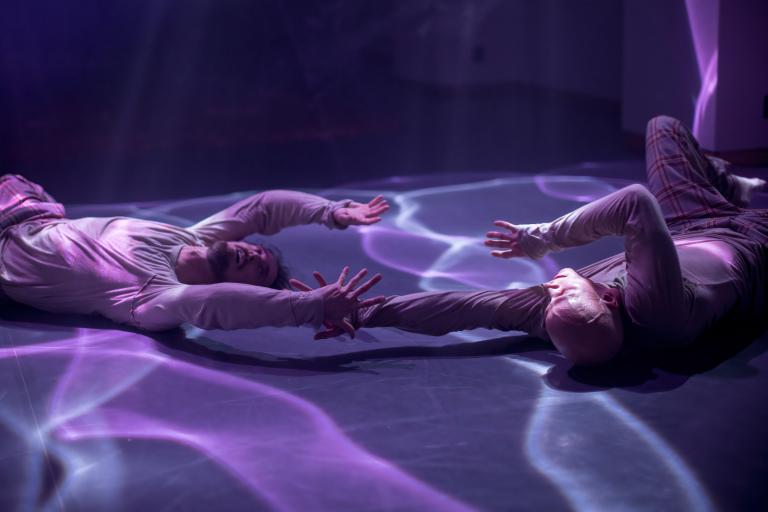 Två dansare ligger på golvet i ett lila ljus med vattenliknande reflektioner. De sträcker sina armar ut mot varandra. 