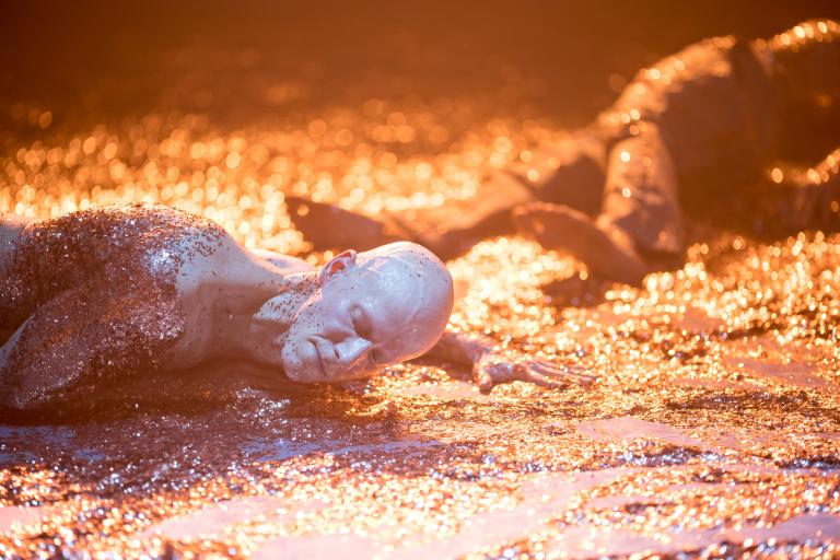 En dansare utan tröja ligger svettig på golvet. Både han och golvet är täckt med rostfärgat glitter.