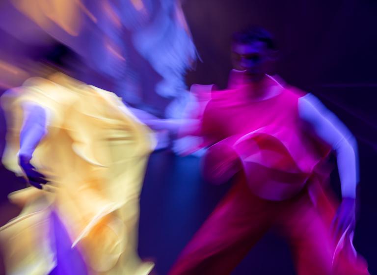 Dansare i ljusgula och skarpt rosa böljande och yviga kläder rör sig i en suddig bild ur föreställningen in:finite