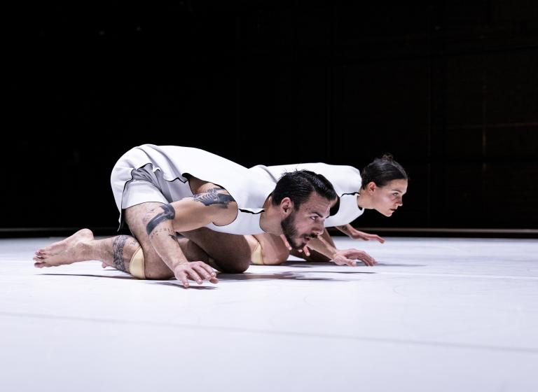 Dansarna Simone Frederick Scacchetti och Amy Josh sitter på knä på ett vitt golv med ansiktena nära golvet och händer och fötter svävande i luften. svävande 