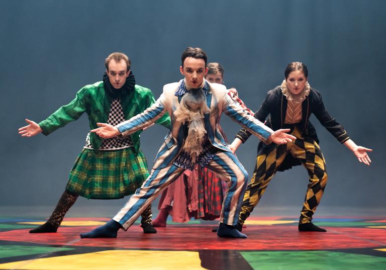 Fyra dansare i färgstarka kostymer med krusiga kragar, kiltar och volangkjolar böjer sig fram mot dig med armarna sträckta utåt och neråt.