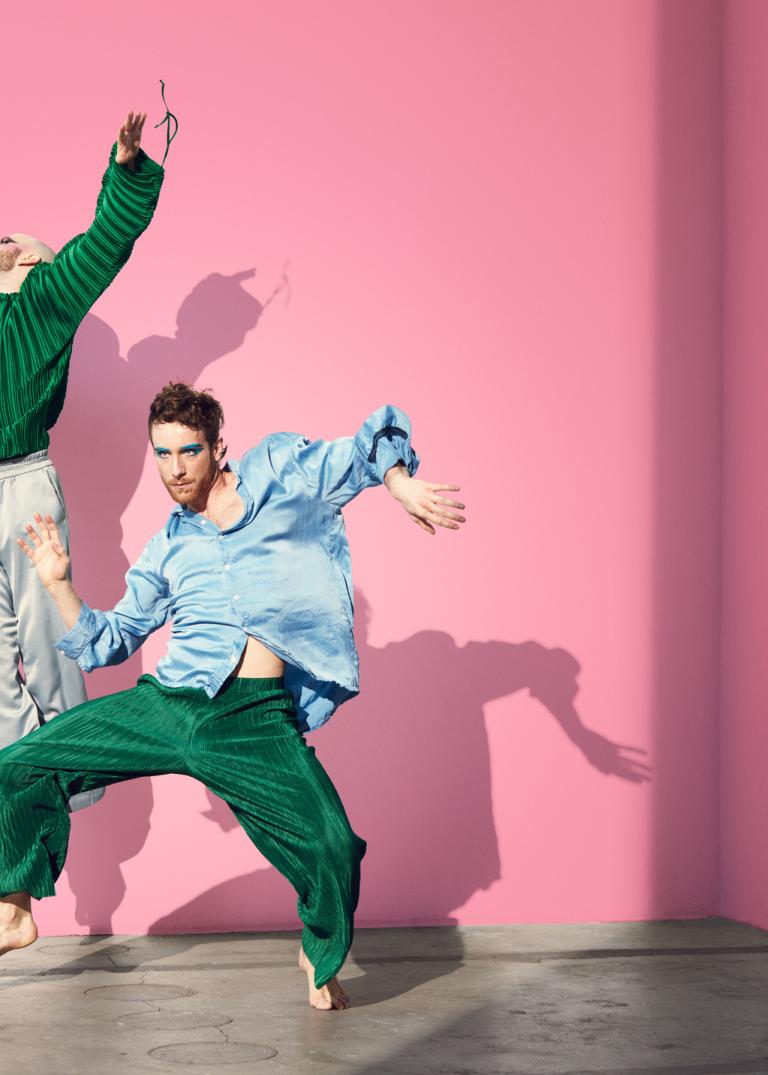 Två dansare i ljusblå och gröna fladdriga kläder dansar framför en rosa vägg.