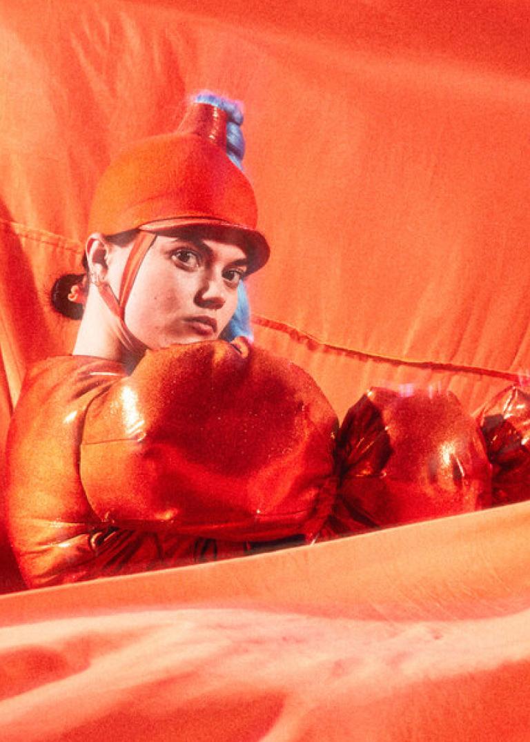 En dansare i orange bubbliga kläder och en ridhjälmsliknande hatt med en lång ljusblå fläta står bland böljande orange tyg framför och bakom henne.