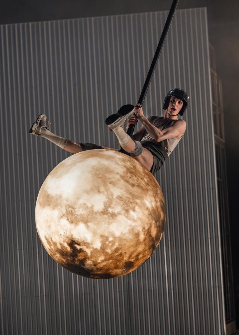 Dansaren Carl Peters sitter på ett stort månliknande klot som hänger högt upp i luften i Skånes Dansteaters jubileumsföreställning Wing Ding. Han har shorts och ett linne och en discobollshjälm på sig och svingar sig med klotet som en gunga.