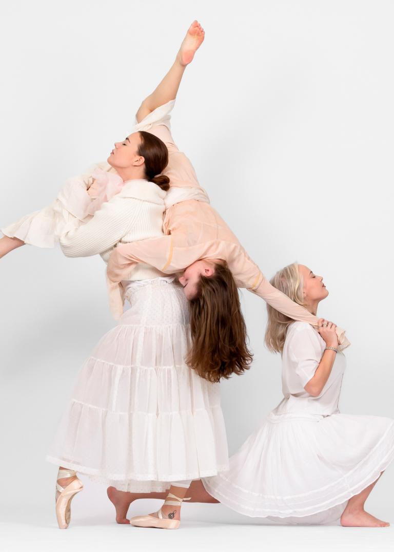 Tre dansare i långa vita kjolar står tillasammans i ett vitt rum. Den ena lyfter den andra över axelm och den tredje sitter på ett knä och håller den andra dansaren i handen.