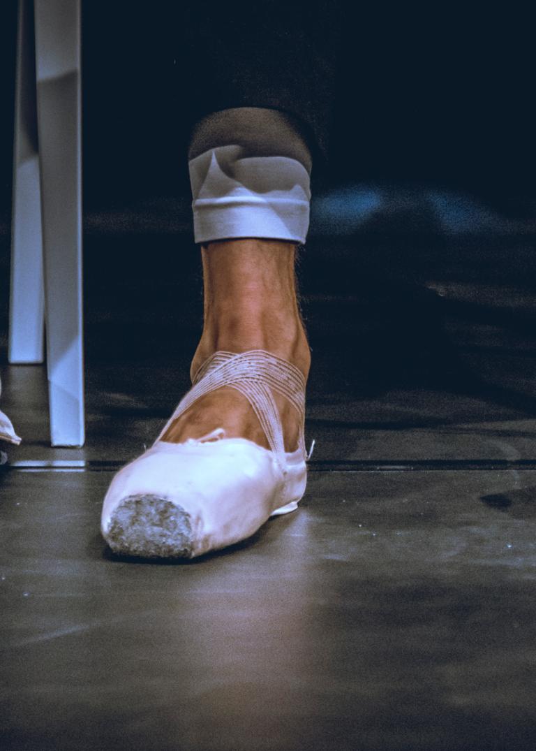 Ett par slitna sneakers står på golvet bredvid en fot i en balettsko.