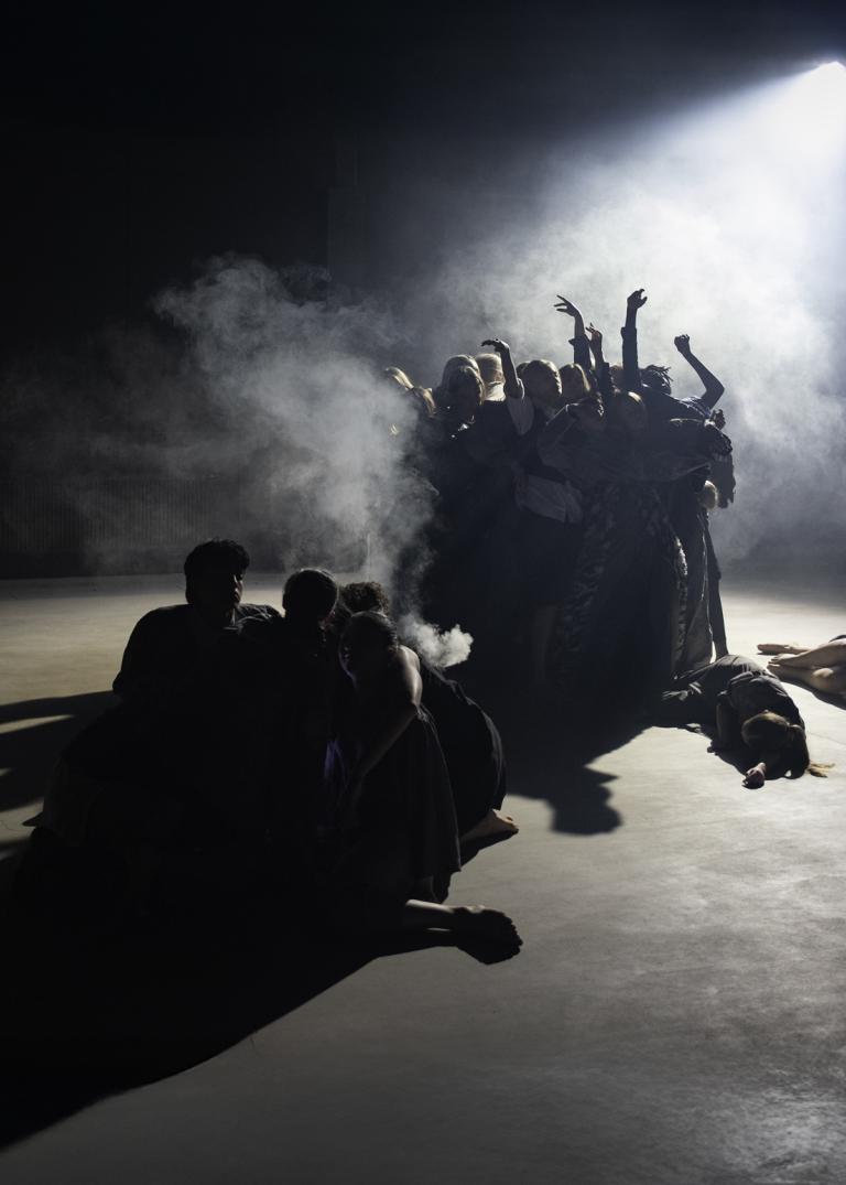 Två grupper av unga dansare rör sig på en scen. En grupp står upp i rökigt ljus från en strålkastare. Den andra gruppen rör sig sittande på golvet i den andra gruppens skugga.