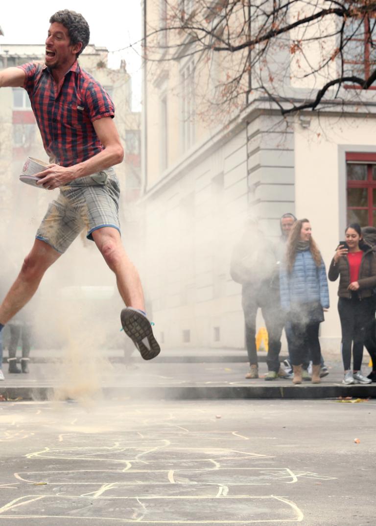 Två dansare i rutiga skjortor och shorts studsar runt på en asfaltyta och kastar dammigt pulver i luften omgivna av publik.