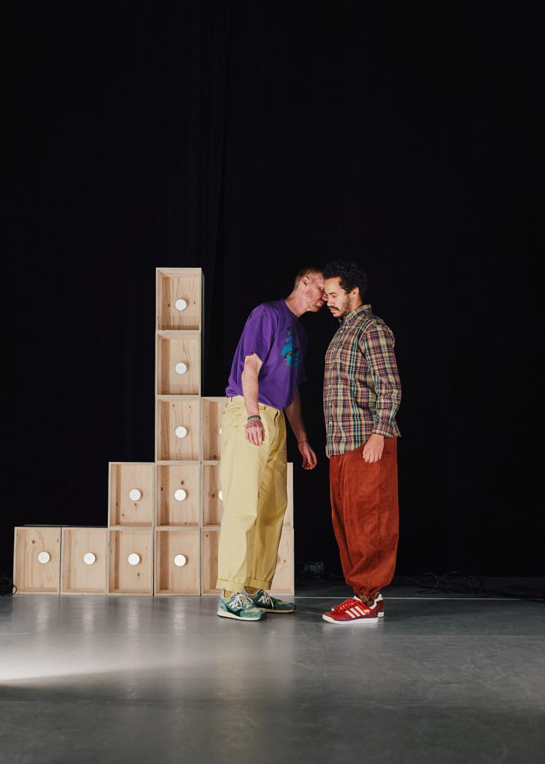 Dansarna Vitkor och Yared står tätt ihop vid staplade omålade träboxar.