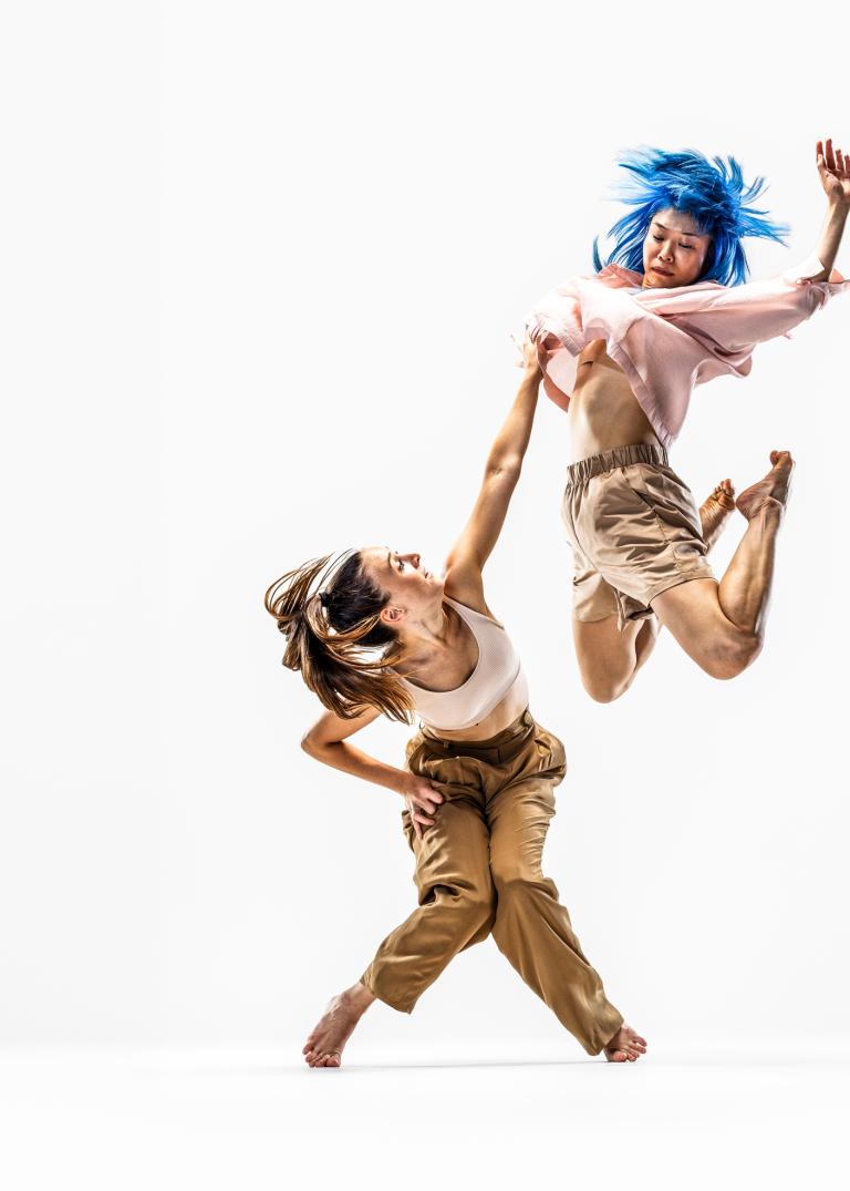 Dansaren Hanna hukar och sträcker sig upp mot dansaren Kat som hoppar högt upp i luften.