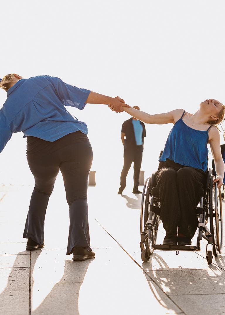 Två dansare hålelr varandra i händerna och lutar bort från varandra i starkt sommarsolljus med havet och andra dansare som bakgrund.
