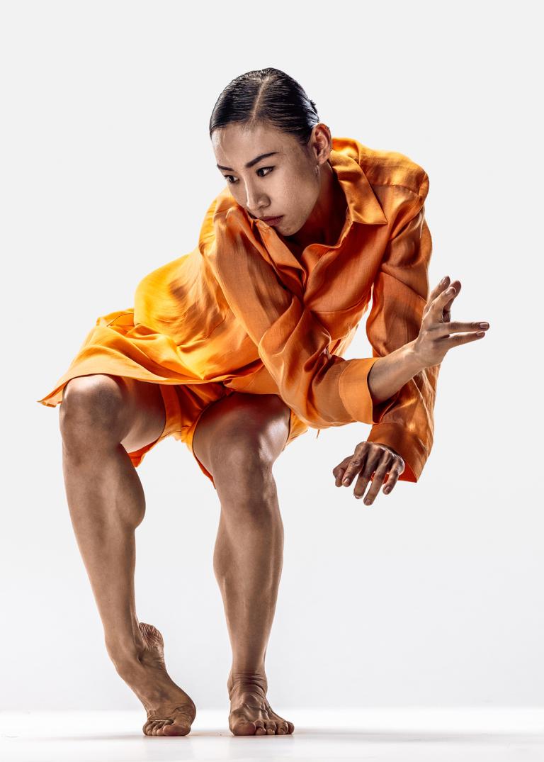 Dansaren Jing Yi Wang klädd i orange shorts och skjorta med svart uppsatt hår dansar med böjda knän och sträcker ut en hand.