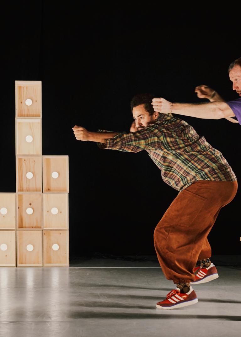 Dansarna Yared och Viktor dansar bredvid varandra bland staplar av omålade trälådor med sladdar och ljuskällor i och håller händerna som om de siktar en pilbåge bortåt. 