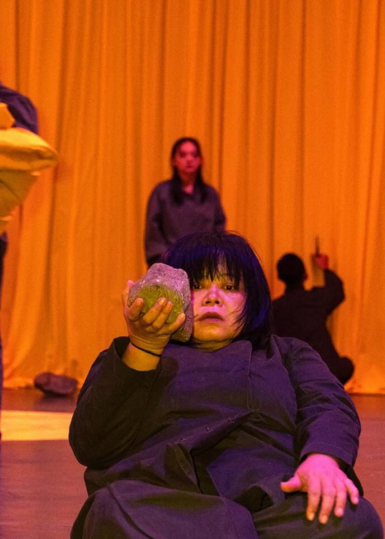 Fyra scenkonstnärer klädda i svarta kläder framför en orange ridå. Melati Suryodarmo halvligger på golvet med en stor sten i ena handen. En konstnär täcks av famnen full av orange kuddar och de två andra konstnärerna står bak i bilden vid ridån. 