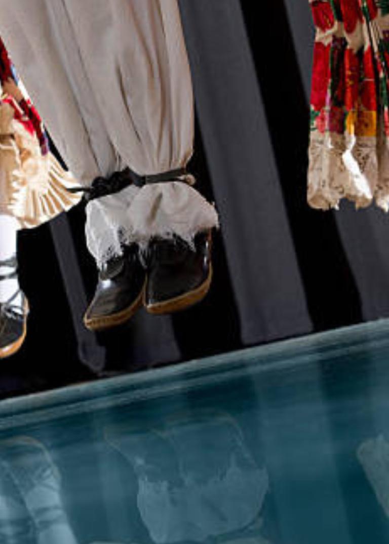 Tre par ben klädda i folkdräktsaktiga byxor och kjolar svävar i luften som om de hoppade upp tillsammans samtidigt.