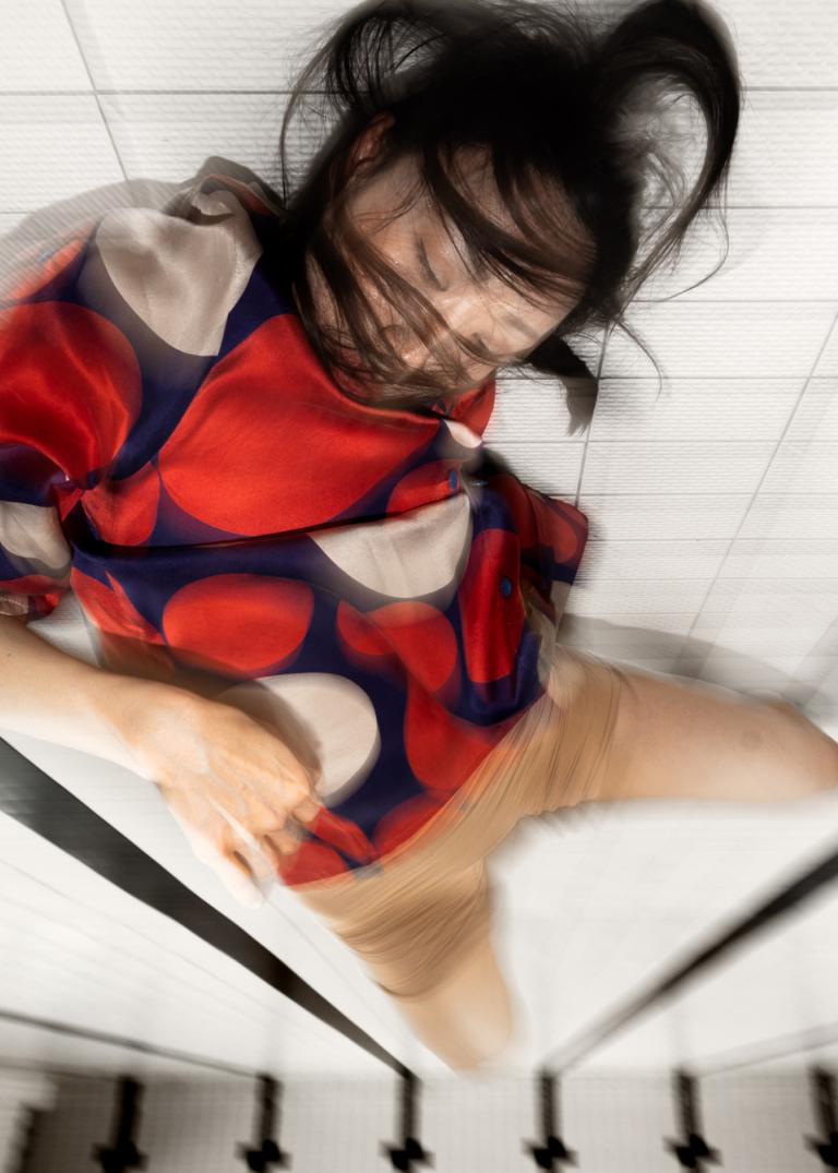 Dansaren Jing Yi Wang i röd och blå kort klänning rör sig på botten av en tömd simbassäng med vitt kakel och långa svarta linjer. Hon blundar och hennes svarta hår sveper runt hennes ansikte.