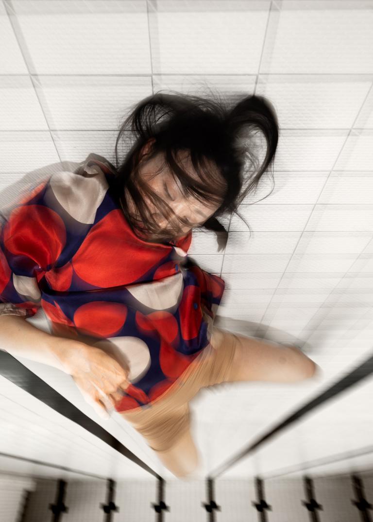 Dansaren Jing Yi Wang i röd och blå kort klänning rör sig på botten av en tömd simbassäng med vitt kakel och långa svarta linjer. Hon blundar och hennes svarta hår sveper runt hennes ansikte.