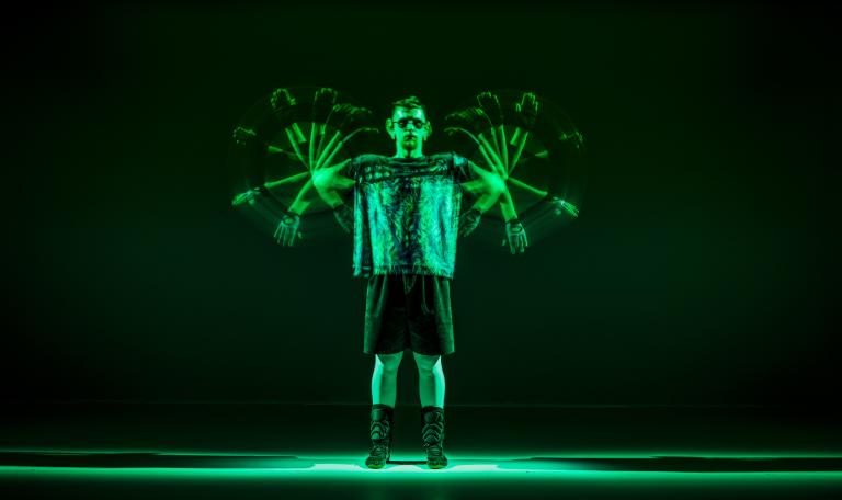 En dansare i mörk t-shirt, short och solglasögon står i grönt ljus och snurrar sina armar likt propellrar.