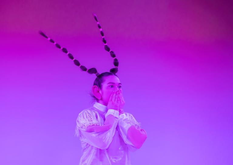 En dansare i ljusa kläder håller sig för munnen. DAnsaren har stora låtsasöron och långt hår i två flätor som står rakt upp bakåt från huvudet.