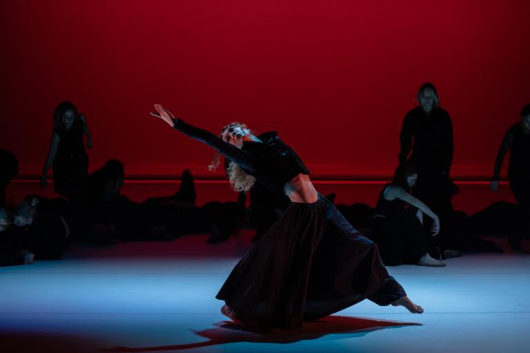 En dansare i lång svart kjol sträcker ut en arm bakåt och tar ett stor steg framåt framför ett antal dansare i mörka kläder framför en röd vägg.