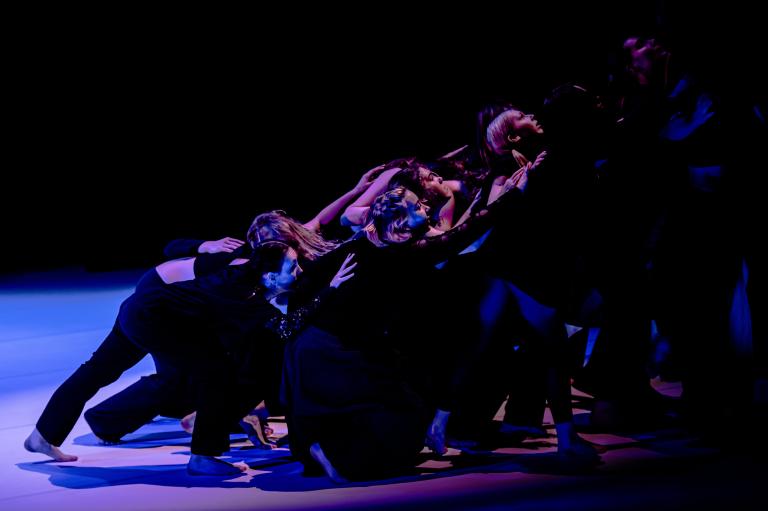 Ett stort antal dansare i svarta kläder lutar sig tillsammans som ett sluttande plan.