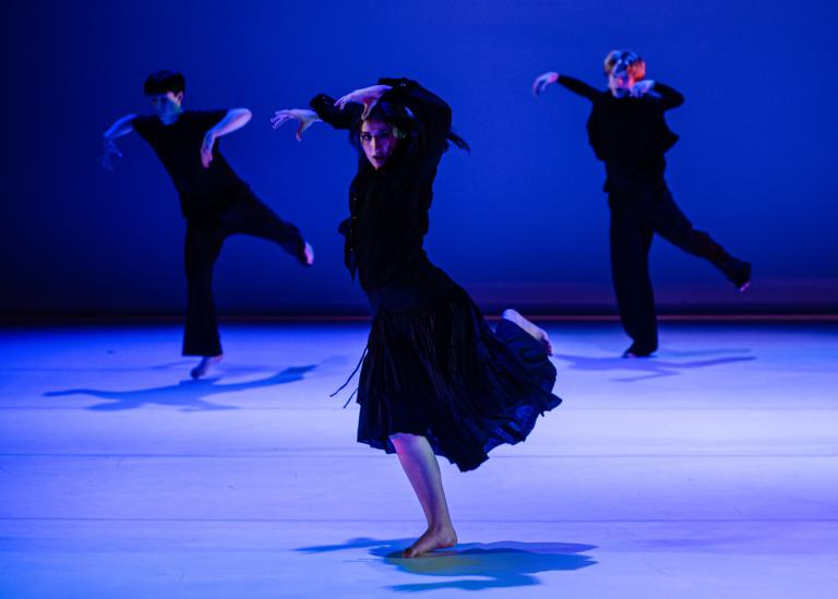 Tre dansare i svart kläder i ett blåskimrande rum dansare med ett ben lyft bakåt och armarna böjda framåt ovanför huvudet.