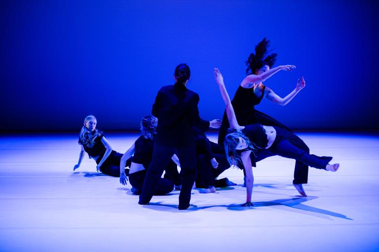 Dansare i svart kläder i ett blåskimrande rum rör sig på olika sätt på golevt och ståendes runt en slags mitt.