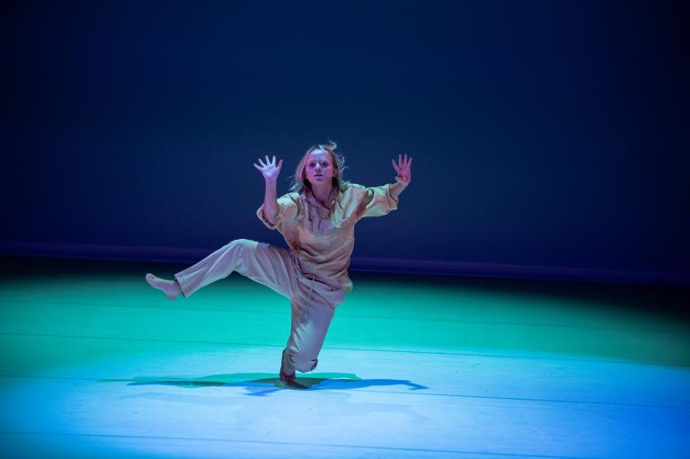En dansare i beiga kläder balanserar på tårna medan hennes andra ben är böjt och lyft upp i luften och hon visar sina handflator.