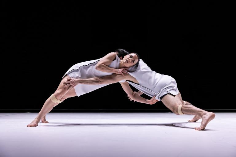 Två dansare i korta vita klänningar lutar sig axel mot axel så att de nästan vilar horisontellt mot ett vitt golv.