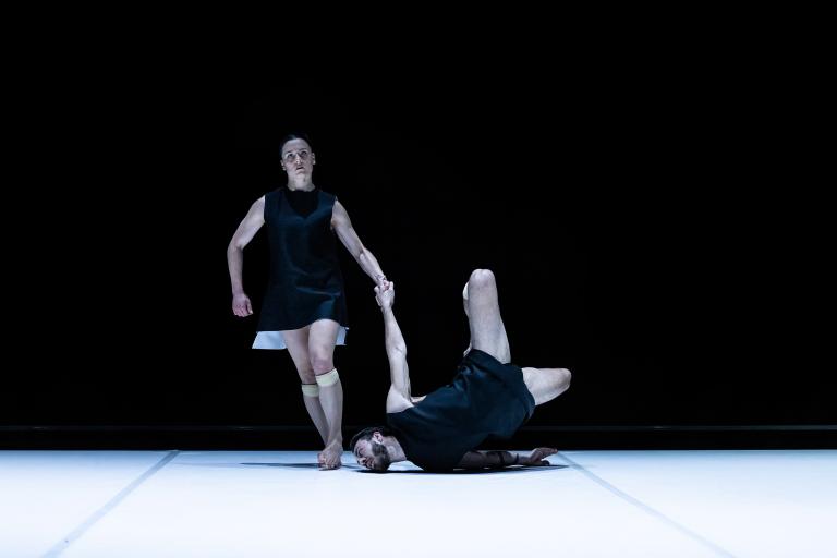 Dansaren Amy Josh går över ett vitt golv. Hon håller dansaren Simone Frederick Scacchetti i handen medan han snurrar längs golvet med ansiktet mot den vita mattan.