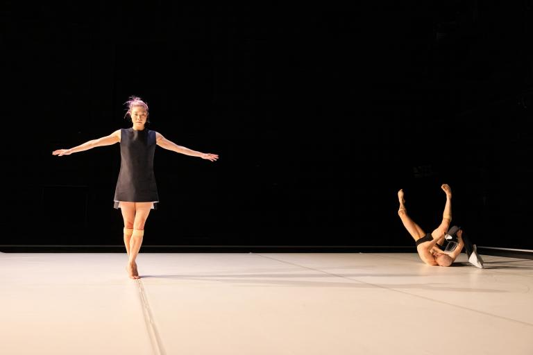 Dansaren Kat Nakui balanserar längs en linje medan dansaren Samuel Denton ligger på rygg med benen uppsträckta i luften i bakgrunden.