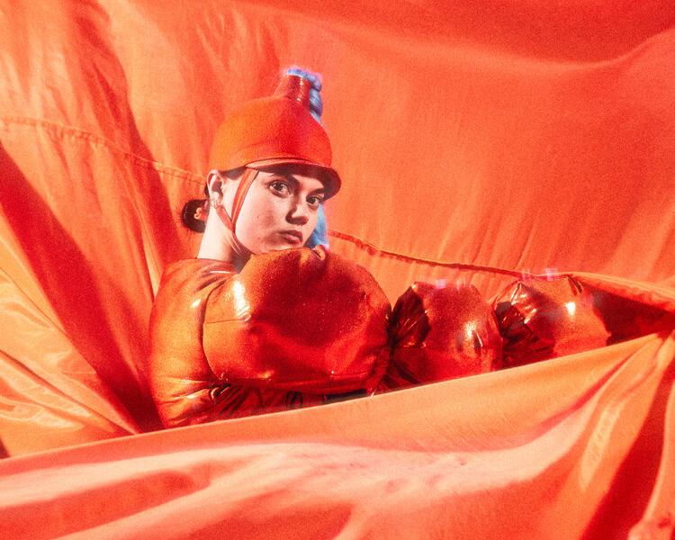 En dansare i orange bubbliga kläder och en ridhjälmsliknande hatt med en lång ljusblå fläta står bland böljande orange tyg framför och bakom henne.