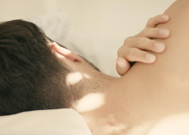 En dansare med mörkt kort hår ligger i en säng med ryggen mot dig och sin hand på sin axel. 
