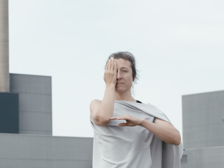 En dansare i grå bljande tunika står framför BArsebäcks kärnkraftverk. Hon håller en hand för ena ögat och skapar en vinkel med sina armar. 