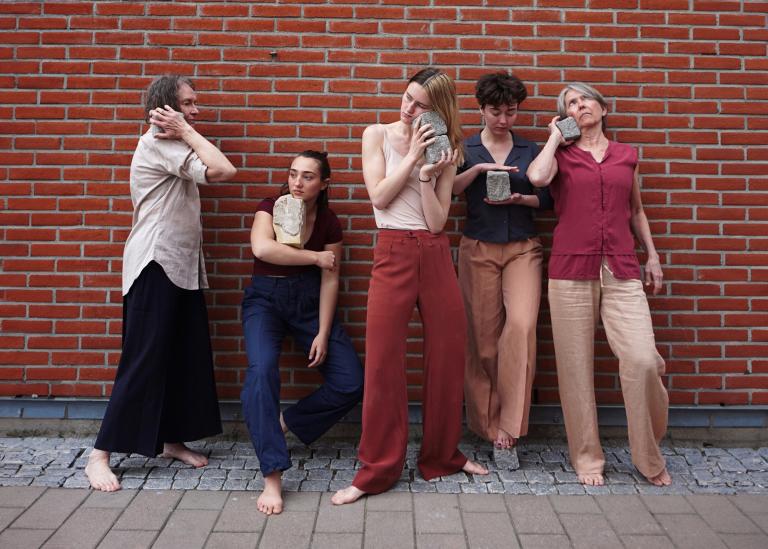 Fem dansare står framför en tegelvägg. De håller varsin kullersten mot olika delar av kroppen.