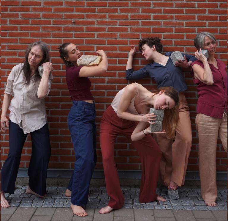 Fem dansare framför en tegelvägg. De håller varsin kullersten mot olika delar av kroppen.