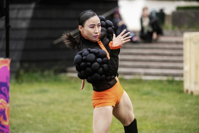 En dansare i svart kavaj med bubbliga axelparti och korta orange shorts.