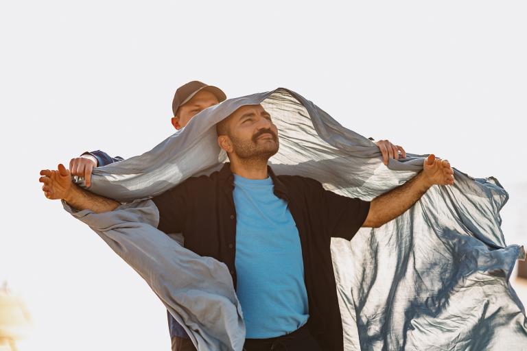En dansare klädd i turkos tshirt och svart skjorta hållar ut sina armar. Bakom honom står en annan dansare och håller ett tunt silverblått tyg som fladdrar runt och mellan dem i Projekt Viaductus föreställning Blå timme.