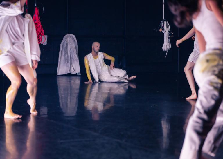 Dansaren Tomás sitter på ett svart golv klädd i vita kläder med gul färg smetad på sig. Framför honom dansar tre dansare i liknande vita kläder. Skånes Dansteaters dansföreställning Attempting Alchemy av koreograf Ludvig Daae.