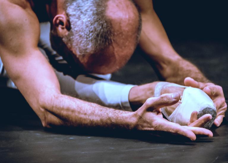 Dansaren Armancio sitter på golvet klädd i balettskor och böjer sig tätt över sitt ben. En del av dansföreställningen The Still Dying Swan.