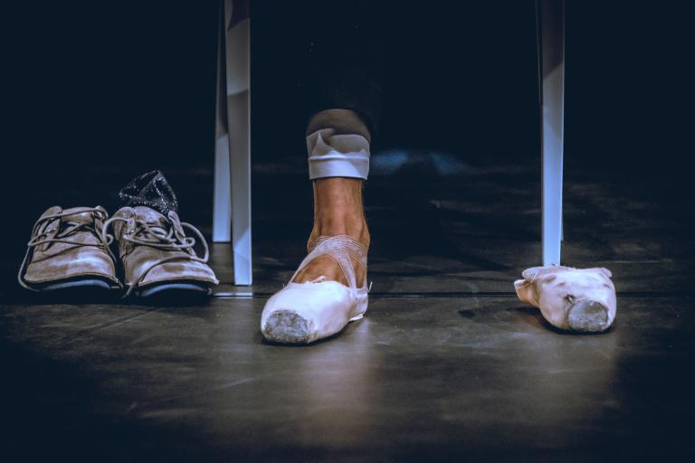 Ett par slitna sneakers står på golvet bredvid en fot i en balettsko.