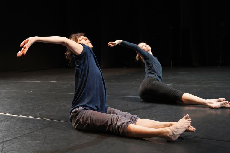 Två dansare i mörka tröjor och byxor sitter på ett mörkt dansgolv. DE sträcker ut sina ben framför sig och sträcker en arm i en båge bakåt. I dansverket The The av koreograferna William Forsythe och Dana Caspersen.
