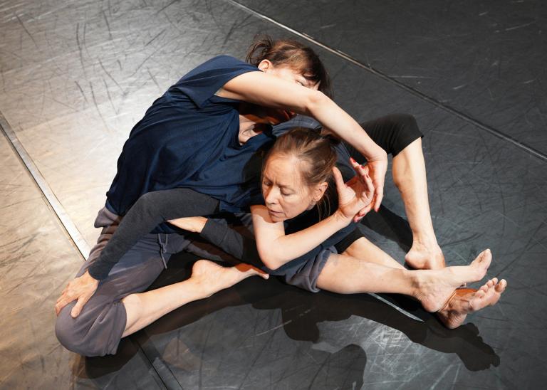 Två dansare i mörka tröjor och byxor sitter på ett mörkt dansgolv. De sträcker sig, sina ben och sina armar om vartannat så att de liknar en knut. I dansverket The The av koreograferna William Forsythe och Dana Caspersen.