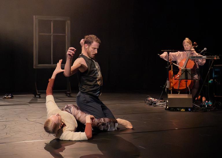 Dansaren Jure sitter på dansaren Anette som ligger på golvet. I bakgrunden spelar Emma Augustsson cello. 
