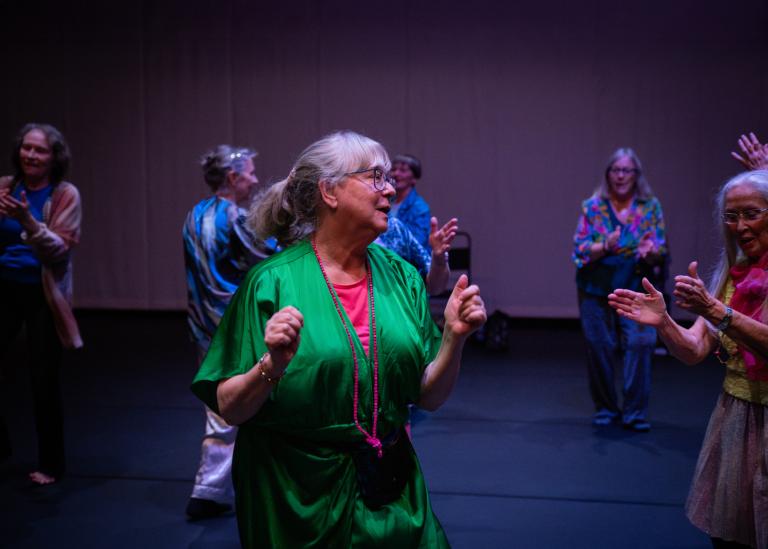 En gästdansare över 60 år med uppsatt hår i hästsvans bär glasögon och är klädd i en grön glansig tunika med en klarrosa top under. De dansar tillsammans med andra dansare i dansföreställningen Jag minns mig själv.