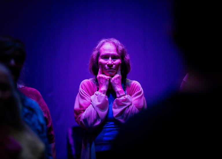 En gästdansare står i lilaskimrande ljus bland andra dansare på scenen i dansföreställningen jag minns mig själv. Hon håller händerna vid hennes käklinje och pekfingrarna vid öronen.